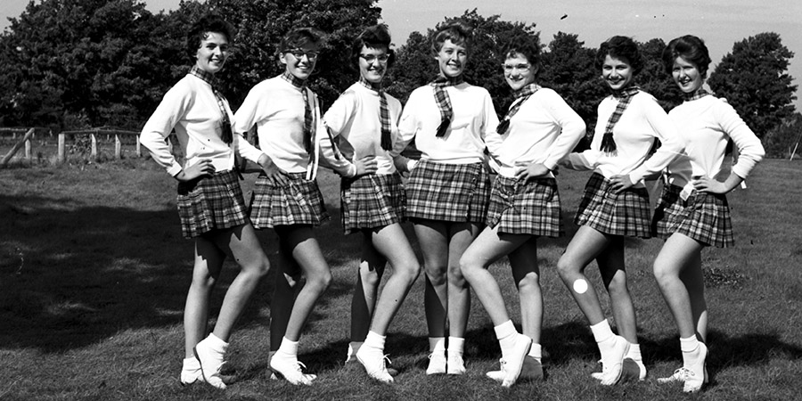 Picture of seven High School cheerleaders in standing poses on a grass field, trees and a fence in the background.