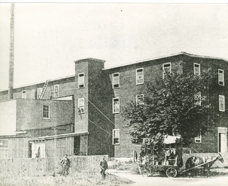 Back and corner of a three-storey factory building, with horse-and-buggy in the foreground.
