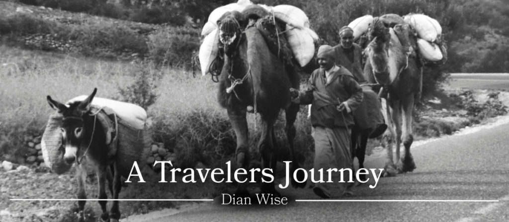 Black-and-white photograph of travelers walking along a rural road with pack animals carrying large bundles. Overlaid text reads “A Travelers Journey” and “Dian Wise.”