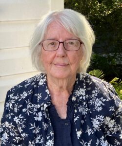 Head-and-shoulders outdoor portrait of a person with short white hair and glasses, wearing a navy-blue shirt and a navy-and-white floral-patterned jacket. The person is standing beside a light-colored wall with green foliage in the background.