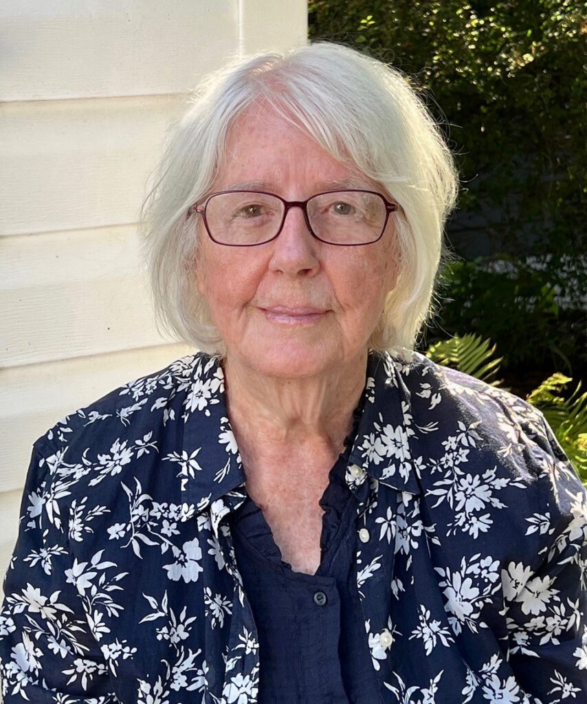Head-and-shoulders outdoor portrait of a person with short white hair and glasses, wearing a navy-blue shirt and a navy-and-white floral-patterned jacket. The person is standing beside a light-colored wall with green foliage in the background.