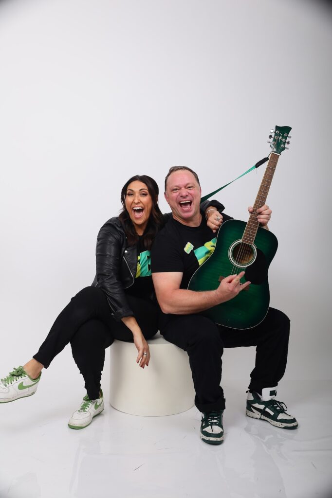 Two people wearing matching black shirts and green-and-white sneakers sit on a white stool in a studio. One person holds a green acoustic guitar while both pose against a plain light-gray background.