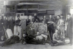 Black and white photo of Group of Gerolamy foundry employees gathered around a fanning mill. inside a workshop with brick walls and wooden shelves. Men wear work clothes and hats, some standing and others reclining