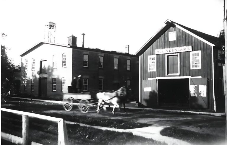 Black and white photograph depicting a horse-drawn carriage moving past two buildings, one of which has a sign reading "W. A. GEROLAMY." The scene captures early 20th-century rural or small-town life, with wooden structures, a fenced area, and a dirt road.