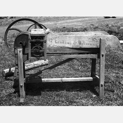 Black and white photograph of an antique wooden and metal corn cutter machine labeled "Gerolamy & Williamson," The machine features visible gears, a large wheel, and a sturdy frame, positioned outdoors on grass with a dirt path in the background.