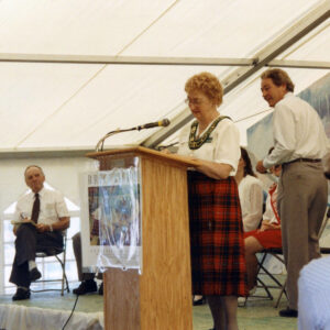 Photograph shows Nellie Johnston delivering remarks at a wooden podium during at IPM opening ceremonies inside a large event tent. Microphone, plaid skirt, seated attendees, standing man, and colorful poster attached to podium provide visual context.