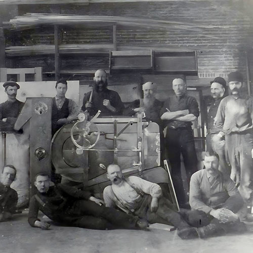Black and white photo of Group of Gerolamy foundry employees gathered around a fanning mill. inside a workshop with brick walls and wooden shelves. Men wear work clothes and hats, some standing and others reclining